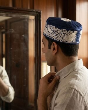 A man in a beige shirt wearing a blue Parsi topi with white embroidery while looking into a mirror.