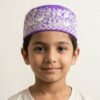 Studio portrait of a smiling young boy wearing a purple Parsi prayer cap featuring intricate white embroidery of birds and floral patterns, set against an off-white background.