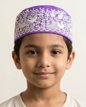 Studio portrait of a smiling young boy wearing a purple Parsi prayer cap featuring intricate white embroidery of birds and floral patterns, set against an off-white background.