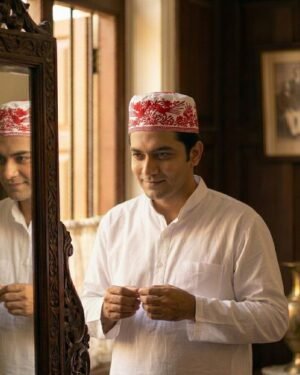 A man in a white tunic wearing a white Parsi topi with red floral embroidery while looking in a mirror.