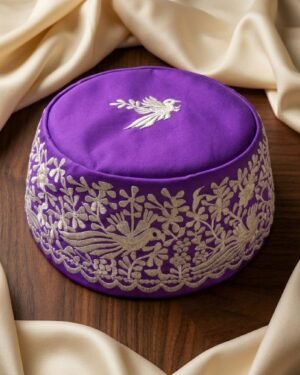 A purple Parsi Gara embroidered topi with a bird motif on the crown, displayed on a wooden surface.