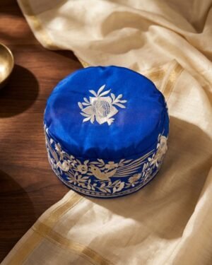 A royal blue Parsi topi with white bird and floral embroidery, displayed on a wooden table with brass bowls .