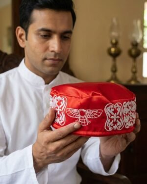 A man wearing a white kurta holds a red embroidered prayer cap with both hands, looking down at it with a contemplative expression