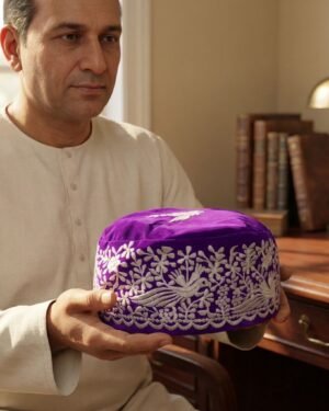 A man wearing a traditional white button-down tunic holds a purple embroidered Parsi cap with both hands in a warm study setting featuring books and a vintage lamp.