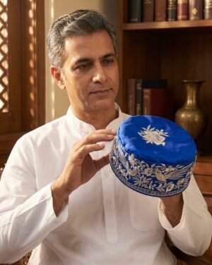 A man wearing a white kurta holds a royal blue Parsi cap with white Gara embroidery, looking at it closely in a traditional room with books and brass vessels.
