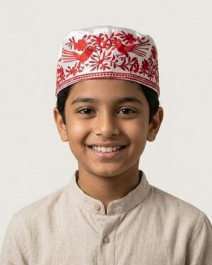 Studio portrait of a smiling young boy wearing a white Parsi prayer cap adorned with intricate red bird and floral Gara embroidery, set against a neutral background.