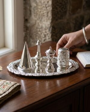 A person’s hand reaching toward a silver Parsi Sesklo set on a wooden table, next to an open book and a traditional embroidered cloth.